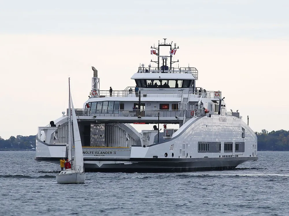 The Wolfe Islander IV ferry crossing the St. Lawrence River between Wolfe Island and Kingston, with passengers on deck and a sailboat passing in the foreground.